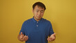 © Krakenimages.com - A handsome young asian man in a blue shirt stands isolated against a vibrant yellow wall, looking pensive and puzzled.