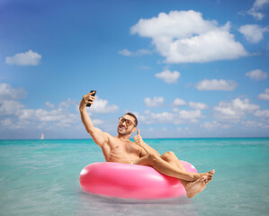 Wall Mural - Man gesturing thumbs up, sitting on a swimming ring and taking a selfie with a smartphone