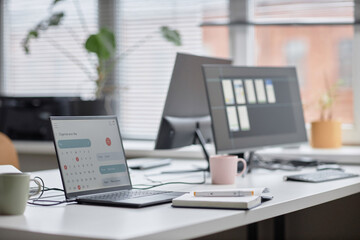 Side view shot in pastel colors of office workplace table with laptop displaying calendar page, notebook and computer monitors in background, focus on screen, copy space