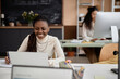 © Seventyfour - Smiling Black woman in office is connecting to remote class or working at laptop. Blackboard on wall in background, copy space