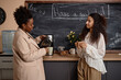 © Seventyfour - Medium long shot of young smiling multiracial students talking while having coffee break pouring coffee in classroom. Multiethnic millennials enjoying drinks against blackboard