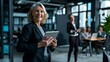 © The Little Hut - Business woman smiling at the camera while holding a tablet pc Mature business woman standing in an office with her colleagues working in the background : Generative AI