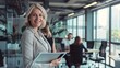 © The Little Hut - Business woman smiling at the camera while holding a tablet pc Mature business woman standing in an office with her colleagues working in the background : Generative AI