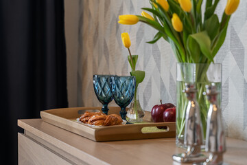 Delicious crispy buns, red apples and two blue glasses on a wooden tray. Yellow tulips in a glass vase. Breakfast in the bedroom