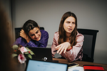 At a wooden table in a contemporary office, two young female workers engage in a lively discussion about project details.