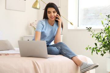 Wall Mural - Female freelancer with laptop talking by mobile phone in bedroom