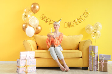 Poster - Happy young woman with birthday cake, gift boxes and decorations sitting on sofa near yellow wall