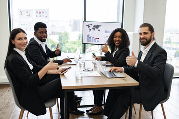 Diverse team of business professionals in a meeting giving thumbs up, showing confidence and success. Positive and productive office environment.