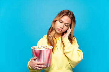 Wall Mural - Teenager girl holding popcorns over isolated blue background with headache