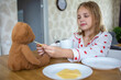 © evgeniia_1010 - A charming blonde baby in cute pajamas with hearts is eating a delicious breakfast and feeding her beloved teddy bear. The concept of proper and balanced nutrition and games for children.