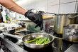 © pavel siamionov - A close-up of a person’s hand in a black glove cooking green beans in a frying pan, with steam rising from pots on a stove in a commercial kitchen.