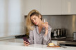 © Volodymyr - Young woman holding a glass of wine in her kitchen. Blond beautiful girl with phone drinking wine. Morning of pretty woman using social media on phone.