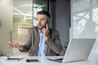 © Liubomir - Businessman in a suit speaking on a phone call and appearing confused while sitting in a modern office ,with a laptop and tablet on the table.