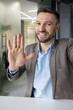 © Liubomir - Smiling businessman waving hand at camera while making video call in a modern office setting, showing cheerful greeting gesture