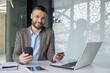 © Liubomir - Smiling businessman sitting at desk with phone and credit card in modern office, showcasing online banking and digital payment.