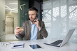 © Liubomir - Worried businessman in a modern office, holding a credit card while talking on the phone and looking at his laptop with concern.
