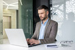© Liubomir - Professional businessman working on a laptop in a modern office setting, focused on his tasks. Background features glass walls and a notebook on the desk.