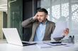 © Liubomir - Stressed businessman holding documents and working on a laptop in a modern office. Man is facing difficulty while analyzing data or dealing with financial problems.