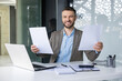 © Liubomir - Confident smiling businessman showcasing documents at his desk in a modern office setting. Professional workspace with laptop and office supplies.