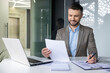 © Liubomir - Smiling businessman working at desk with laptop, reviewing documents and taking notes in a modern office
