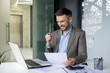 © Liubomir - Smiling businessman working on laptop, analyzing documents in a modern office setting. Celebrating success and achievement.