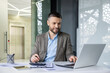 © Liubomir - Smiling businessman working on laptop in modern office, analyzing documents and taking notes. Professional and focused in corporate environment.
