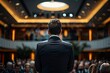 © At My Hat - A man stands in front of a crowd, giving a speech. The audience is attentive and engaged, listening to the man's words. The atmosphere is serious and formal, as the man is dressed in a suit and tie