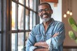 © Bruno Mazzetti - Confident African American man with glasses and a denim shirt standing near a sunlit window, smiling warmly while crossing arms in a cozy indoor setting.