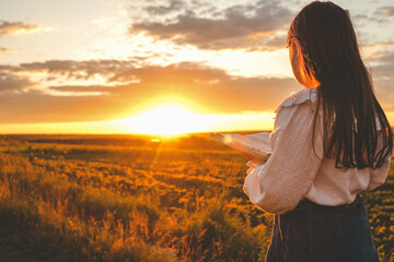 Wall Mural - Girl with a Bible in her hands on a field at sunset, prayer and worship