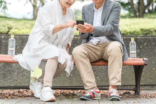 公園のベンチでスマホを見ながら会話する高齢者夫婦（シニア）
