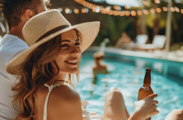 Canvas Print - A happy, smiling couple enjoying drinks by the pool in summer; the woman is wearing an elegant sun dress and hat while holding a drink bottle with her legs crossed on top of each other