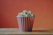 © Mamstock - A bucket of popcorn is positioned on top of a wooden table, with no one around. The popcorn looks freshly popped, and the table is plain in design.