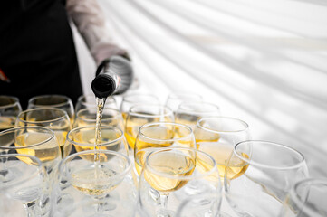 A person in a black sleeve pours a clear, golden liquid into elegant glasses arranged on a table with a white draped background