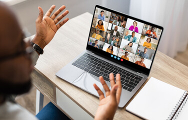 Wall Mural - Black man sits at a desk in front of a laptop screen, participating in a video conference meeting. He has both hands raised, gesturing to the other participants in the call.