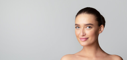 A young woman is smiling while standing in front of a plain gray background. Her expression is cheerful and relaxed as she looks directly at the camera, panorama with copy space