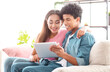 © Prostock-studio - African American teen couple is sitting on a couch in their living room. They are both smiling and looking at a tablet. They appear to be enjoying each others company and the content on the tablet.