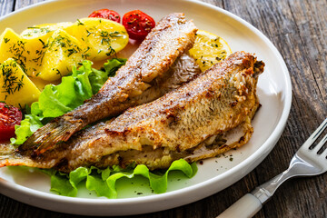 Wall Mural - Fried sea bass served on lettuce with boiled potatoes and  lemon on white plate on wooden table
