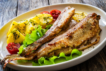 Poster - Fried sea bass served on lettuce with boiled potatoes and  lemon on white plate on wooden table

