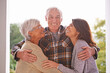 © Marius Venter/peopleimages.com - Outdoor, portrait and daughter hug old parents, visit and love on patio of home, smile and happiness. Family, man and women embrace with care, girl and mother with father, retirement and house