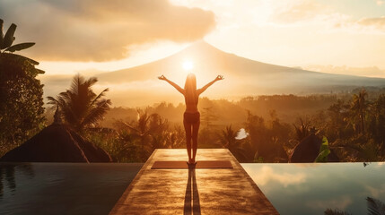 Wall Mural - Woman practicing yoga at sunrise with tropical landscape and mountains in the background, standing on a yoga mat next to an infinity pool.