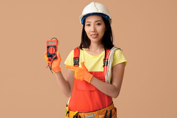 Young female electrician pointing at multimeter on beige background