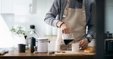 Asian man in apron pouring coffee in cup while standing near cooking surface in kitchen