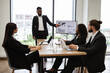 © sofiko14 - Diverse business team in formal attire having a meeting. Professional man presenting data on a screen while colleagues listen attentively. Modern office setting with city view in background
