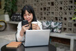 © bongkarn - A woman is sipping coffee and watching videos on her phone while working remotely from a coffee shop