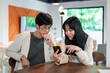 © bongkarn - Two Asian friends are looking at a phone screen and talking while sitting together in a coffee shop.