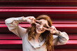 © Westend61 - Happy woman standing and posing peace sign in front of eyes near corrugated iron