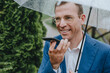 © Westend61 - Smiling businessman holding umbrella and talking through speaker phone
