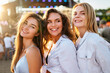 © artiemedvedev - Three smiling young women enjoy summer music festival on sandy shore. Friends pose in sunlight, stage lights behind. Casual fashion, fun, togetherness at outdoor concert event by sea.