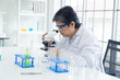 © Rakchanok - Asian senior female scientist wearing safety goggles and a white lab coat examining chemical samples using a microscope in modern laboratory, various lab equipment and test tubes are arranged on table