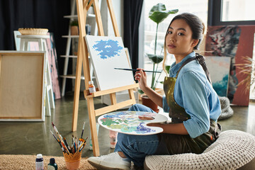 Wall Mural - A young Asian woman in an apron sits in her art studio, painting on a canvas.